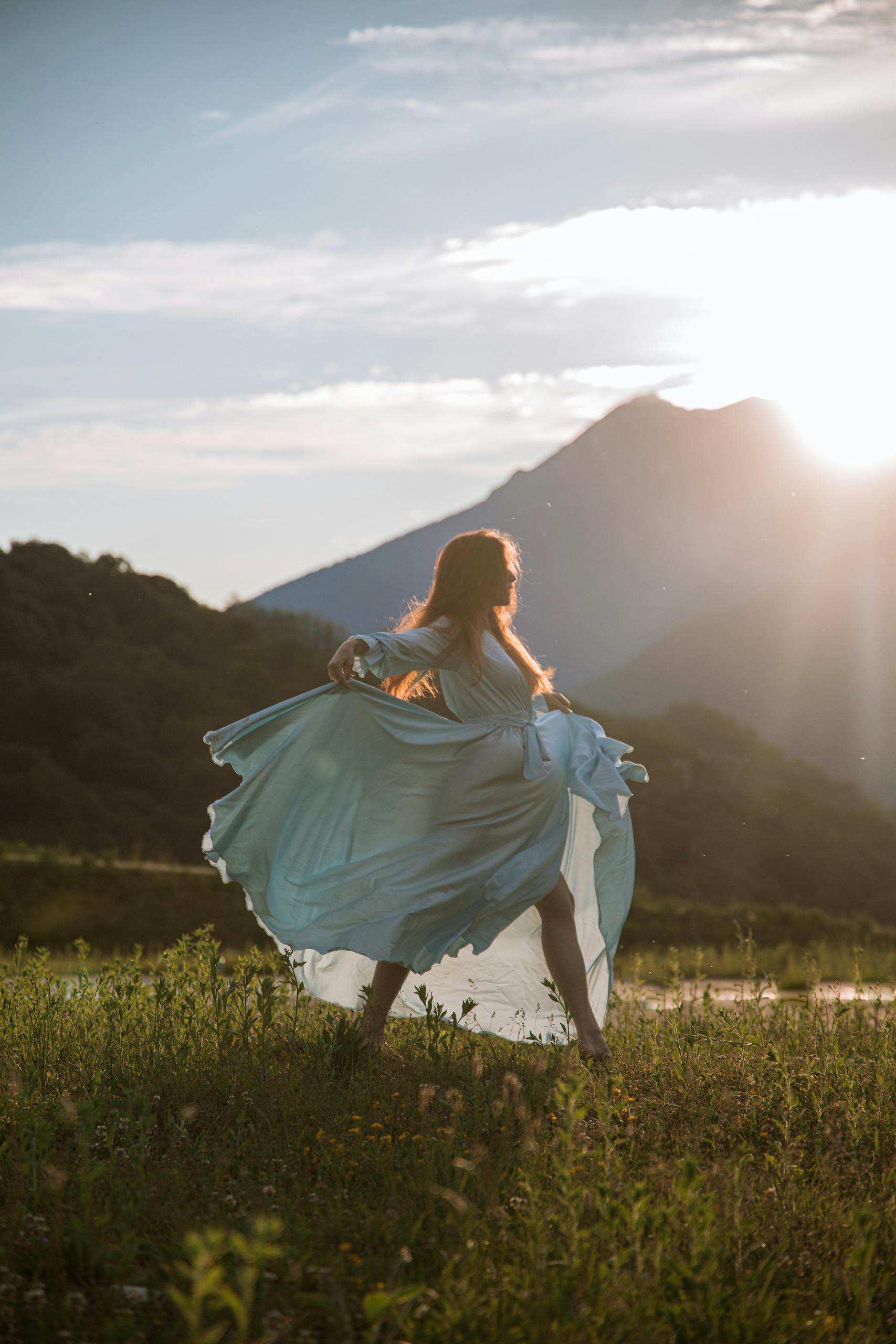 A woman in a flowing blue dress dances gracefully at sunset with mountains in the background.